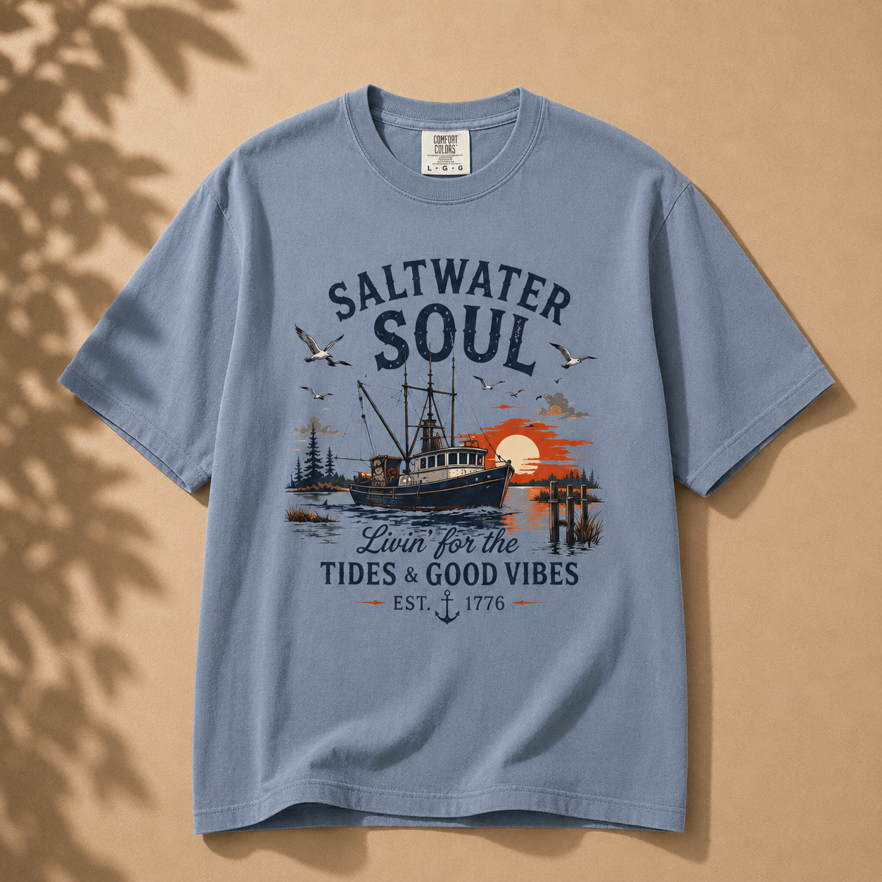 A light blue crewneck t-shirt with a vintage-style nautical graphic reading 'SALTWATER SOUL' and 'Lawn for the Tides & Good Vibes EST. 1776', featuring a fishing boat, seagulls, and a sunset, displayed flat against a warm beige background with soft leaf shadows.