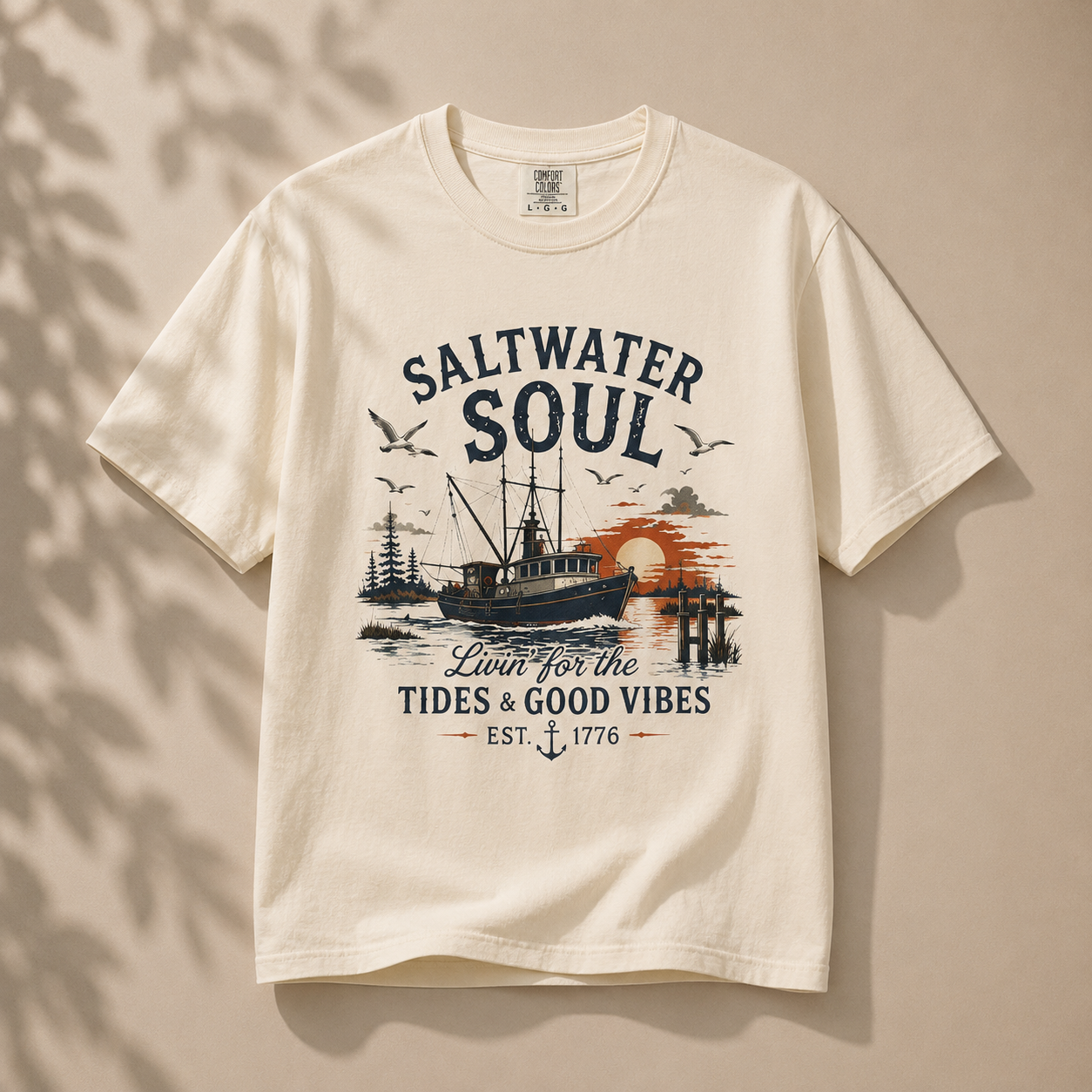 An off-white short-sleeve t-shirt with a vintage-style 'Saltwater Soul' nautical graphic, displayed against a warm beige background with soft leaf shadows.