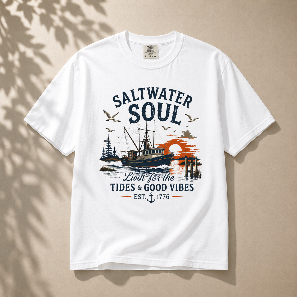 A white short-sleeve crewneck t-shirt with a vintage-style nautical graphic on the front. The graphic features the text 'SALTWATER SOUL' in dark blue, a fishing boat, seagulls, a sunset in orange, and the phrase 'Lift for the Tides & Good Vibes EST.