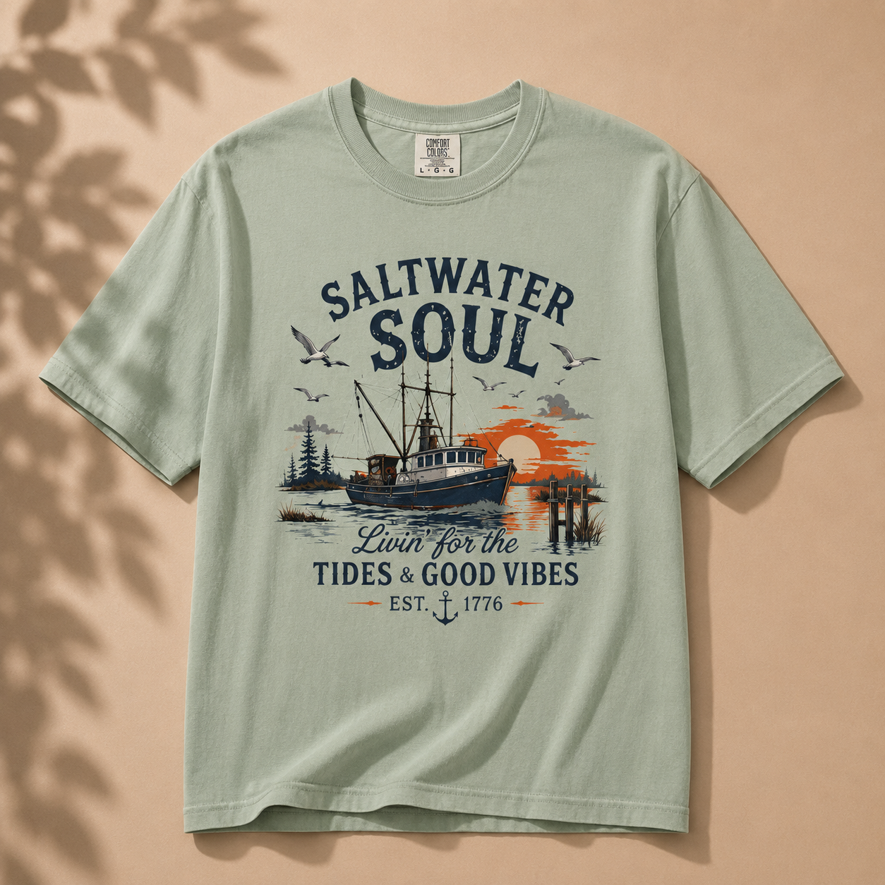 A sage green short-sleeve t-shirt with a vintage-style fishing boat graphic and the words 'SALTWATER SOUL' and 'Livin for the TIDES & GOOD VIBES' on a warm beige background with soft leaf shadows.