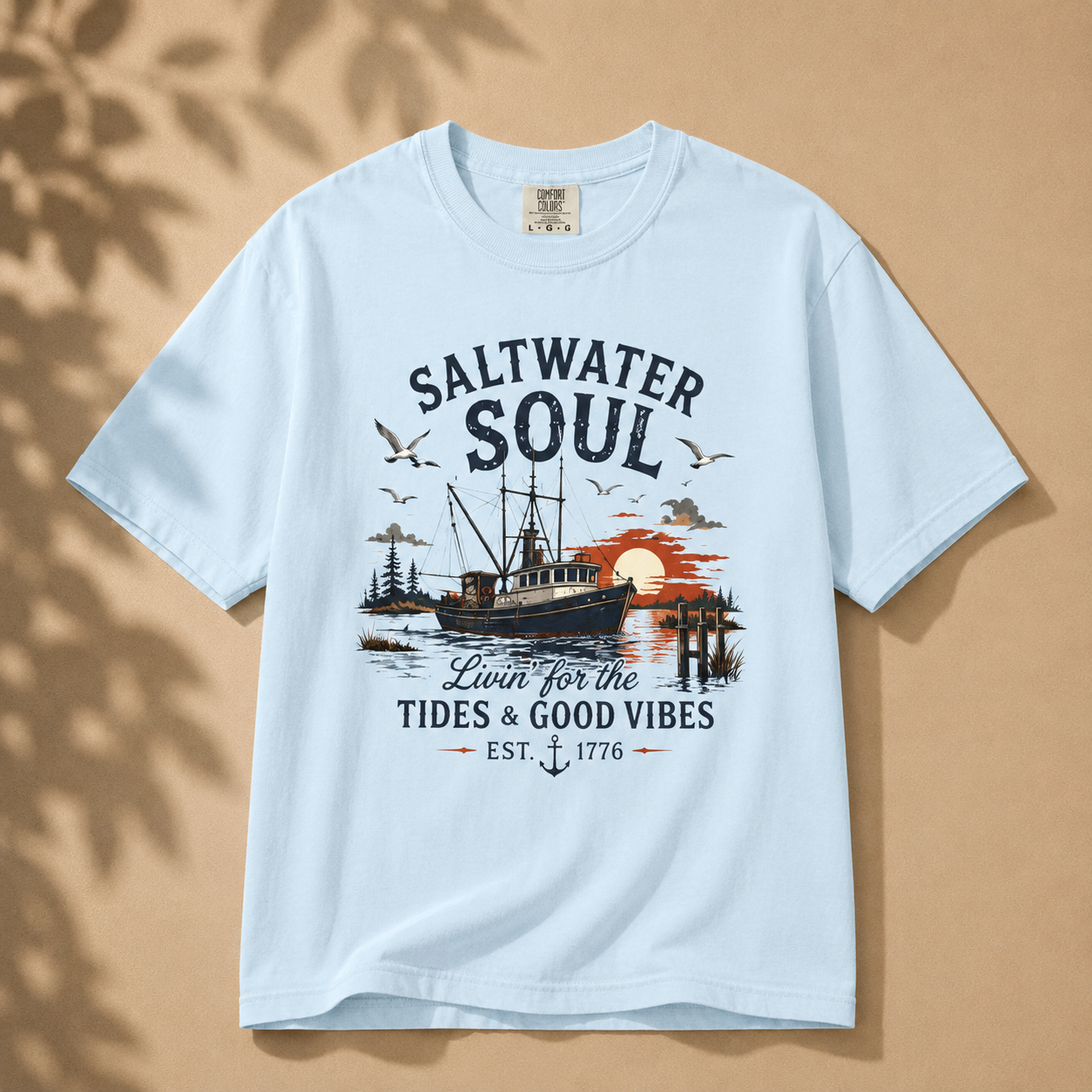 A light blue short-sleeve graphic t-shirt laid flat on a warm beige background with soft leaf shadows. The shirt features a central graphic of a fishing boat, birds, a sunset, and the text 'SALTWATER SOUL' and 'Loved for the TIDES & GOOD VIBES EST.