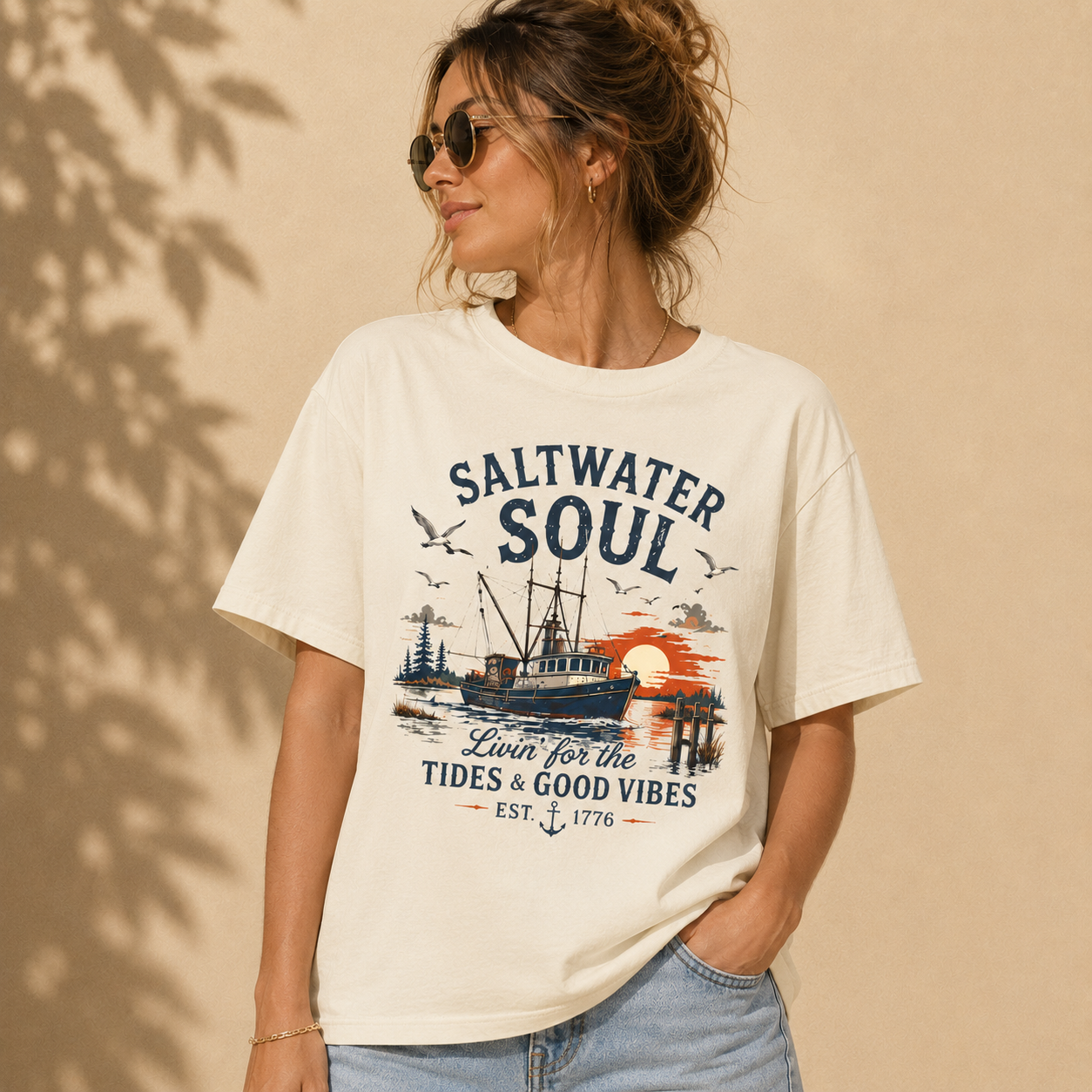 A woman wearing a cream-colored graphic t-shirt with a 'Saltwater Soul' nautical print, paired with light blue jeans, set against a warm beige background with soft leaf shadows.