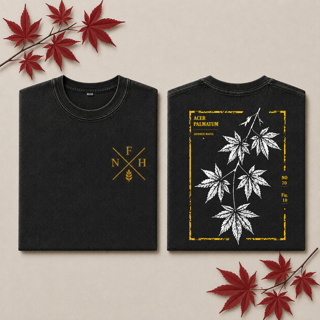 Two folded black T-shirts shown on a beige background with red maple leaves as props. The left shirt displays a gold crossed letters and wheat emblem on the chest, and the right shirt shows a back design with white maple leaves inside a distressed gold border and the text 'ACER PALMATUM'.