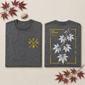 Two folded black graphic T-shirts displayed side by side on a light textured surface. The left shirt shows a large white fern leaf design with a gold rectangular border and text reading 'PTERIDIUM AQUELUM' and 'COLDWATER FERN'.