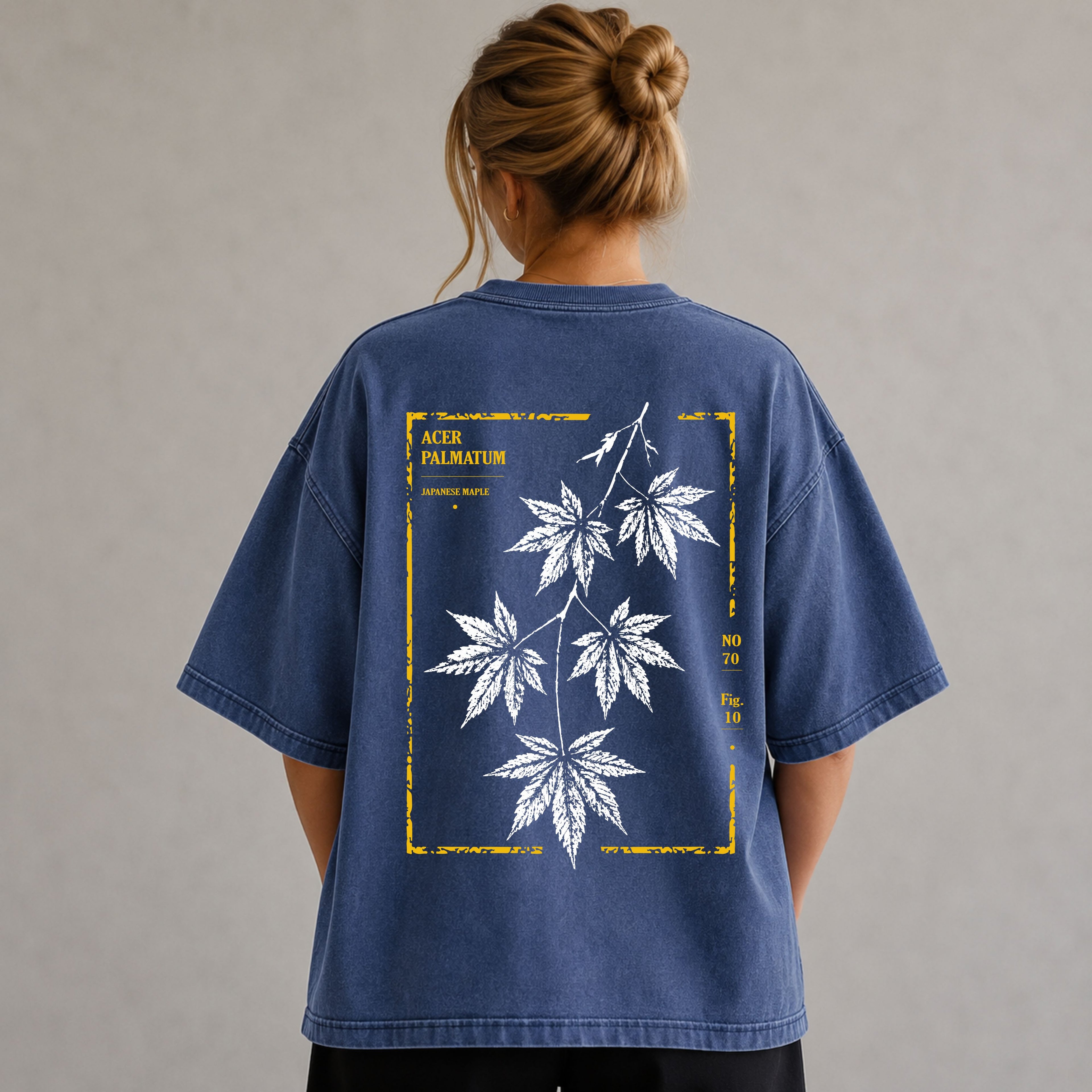 A woman viewed from behind wearing an oversized blue graphic T-shirt with a large white maple leaf print and yellow text on the back, set against a neutral gray background.