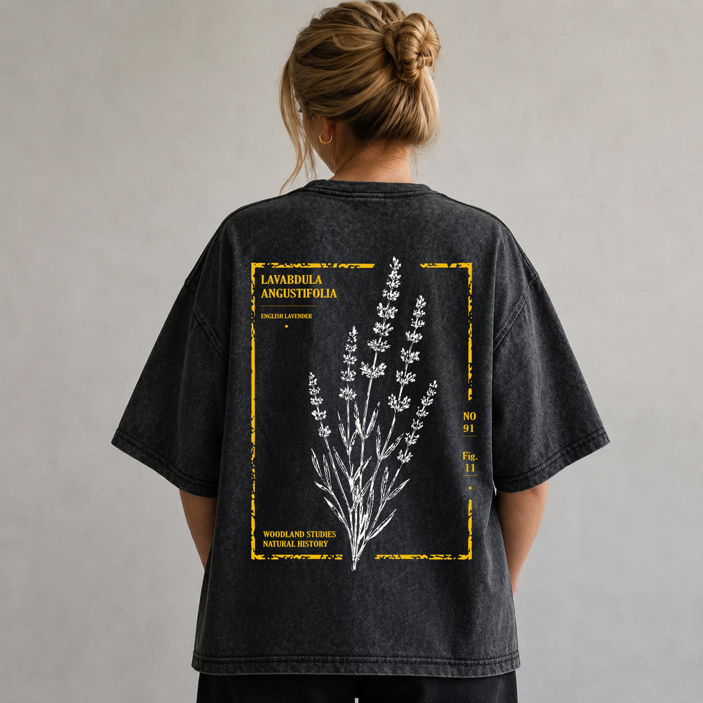 A person viewed from behind wearing an oversized dark gray washed T-shirt with a large botanical graphic on the back. The design shows white lavender sprigs inside a distressed yellow border with text including 'Lavandula Angustifolia' and 'Woodland Studios Natural History.