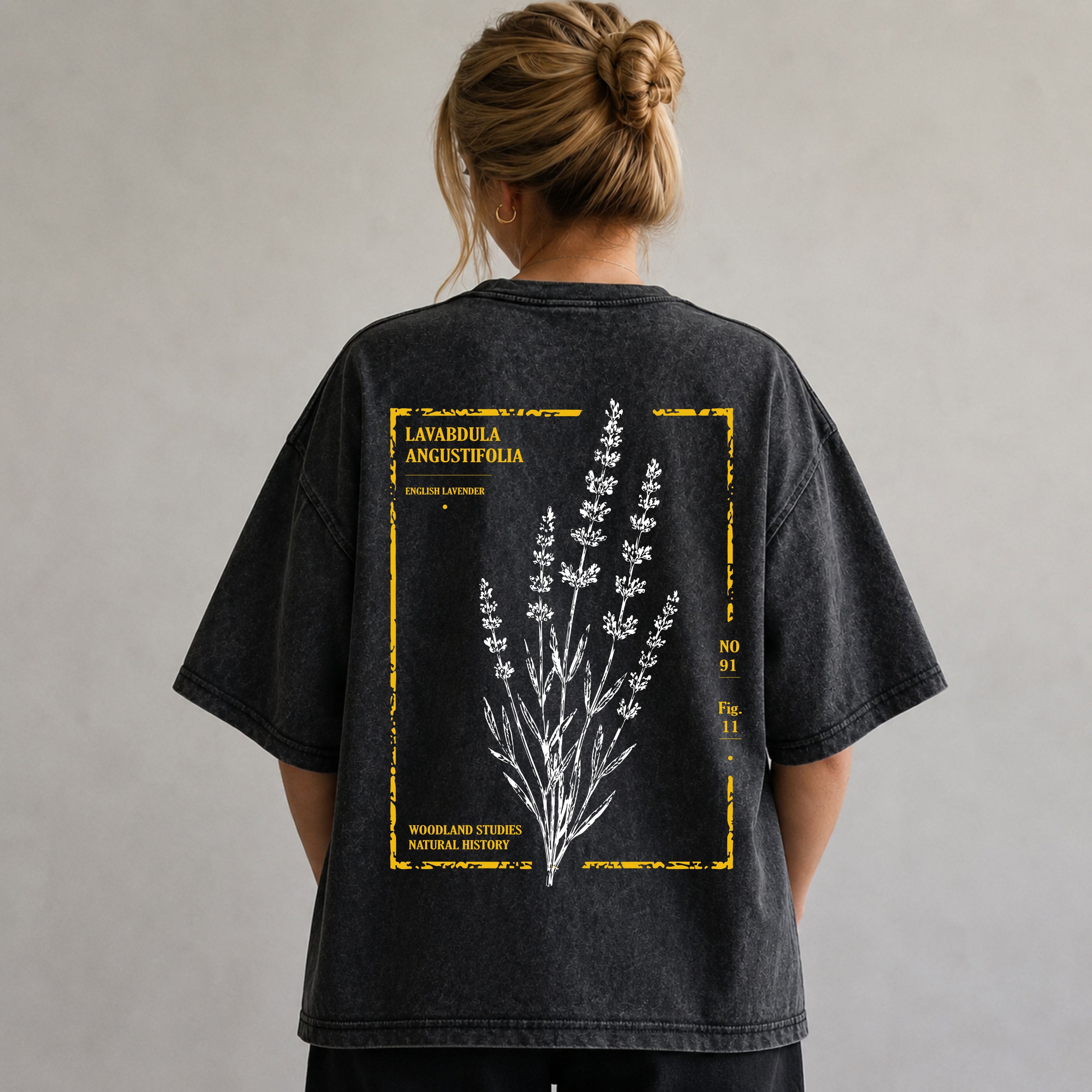A person viewed from behind wearing an oversized dark gray washed T-shirt with a large botanical graphic on the back. The design shows white lavender sprigs inside a distressed yellow border with text including 'Lavandula Angustifolia' and 'Woodland Studios Natural History.