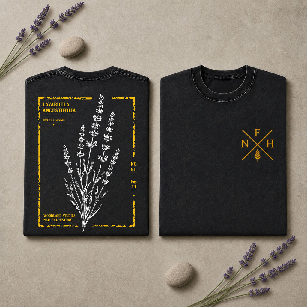 Two black short-sleeve T-shirts laid flat on a beige surface with lavender sprigs and small stones. The left shirt shows a white botanical lavender illustration with yellow text and border details, and the right shirt has a small yellow monogram-style logo on the chest.