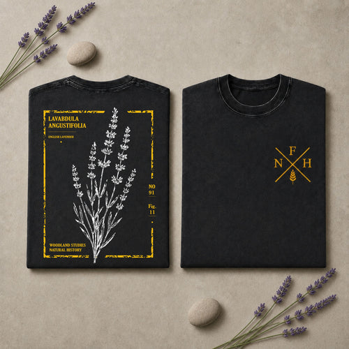 Two black short-sleeve T-shirts laid flat on a beige surface with lavender sprigs and small stones. The left shirt shows a white botanical lavender illustration with yellow text and border details, and the right shirt has a small yellow monogram-style logo on the chest.