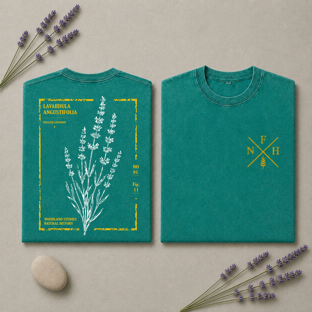 Two views of a teal short-sleeve T-shirt laid flat on a beige surface with lavender sprigs and a stone. The left view shows a large white botanical illustration with yellow text reading 'LAVANDELA ANGUSTIFOLIA' and small yellow details.