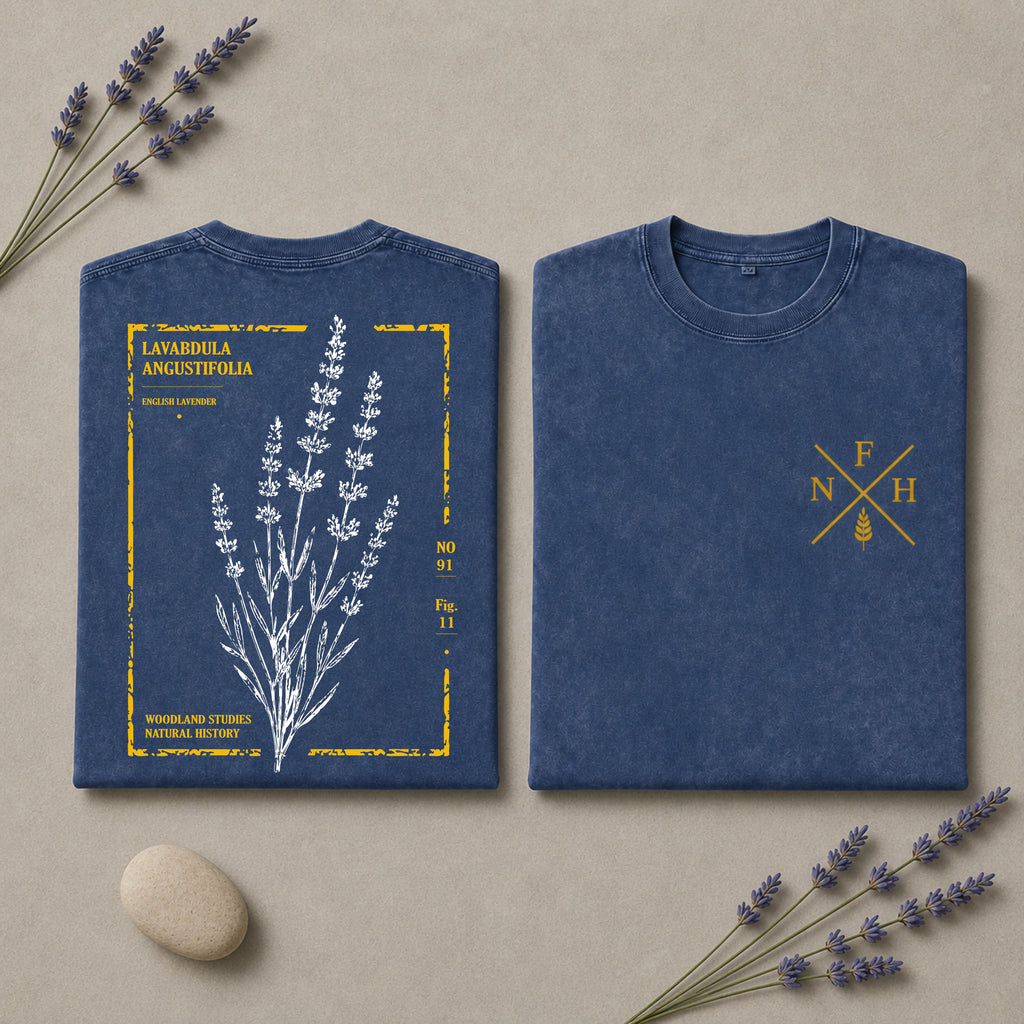 Two folded blue T-shirts displayed on a beige surface with lavender sprigs and a small stone. One shirt shows a front botanical print of lavender with yellow text and border details, and the other shows a small yellow crossed-letter chest graphic.