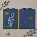 Two folded blue T-shirts displayed on a beige surface with lavender sprigs and a small stone. One shirt shows a front botanical print of lavender with yellow text and border details, and the other shows a small yellow crossed-letter chest graphic.