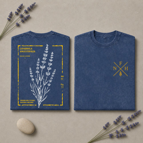 Two folded blue T-shirts displayed on a beige surface with lavender sprigs and a small stone. One shirt shows a front botanical print of lavender with yellow text and border details, and the other shows a small yellow crossed-letter chest graphic.