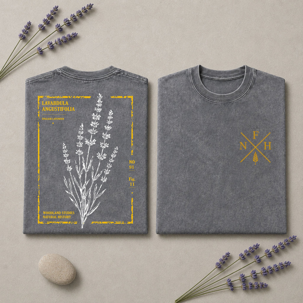 Two folded gray T-shirts displayed side by side on a beige surface with lavender sprigs and a small stone. The left shirt shows the back with a white lavender illustration and yellow text reading 'Lavandula Angustifolia' and 'Moorland Studs Organic Farm.