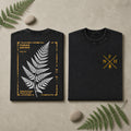 Two folded black T-shirts displayed side by side on a beige surface with decorative fern leaves and stones. The left shirt shows a large white fern leaf graphic framed by distressed yellow text and border details.