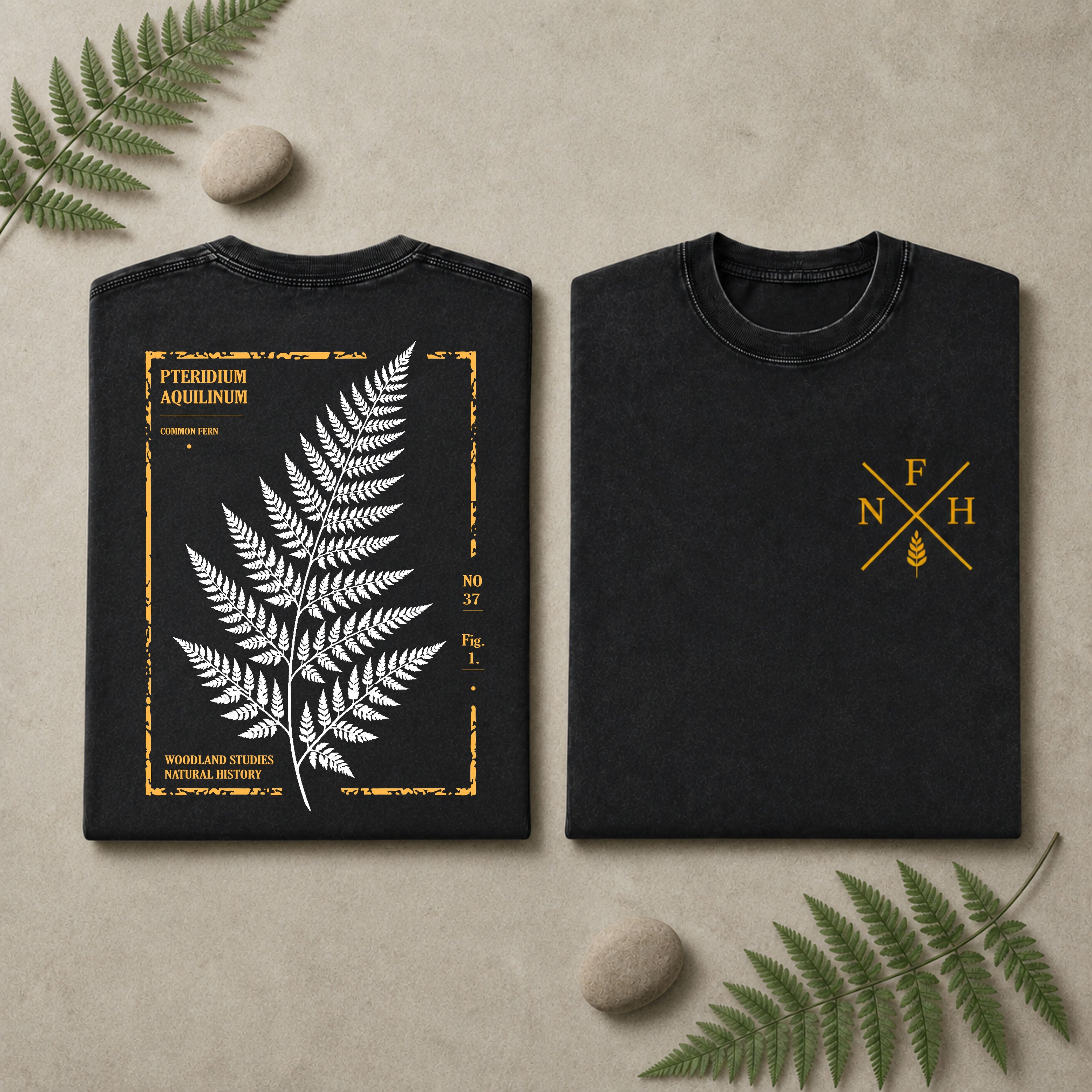 Two folded black T-shirts displayed side by side on a beige surface with decorative fern leaves and stones. The left shirt shows a large white fern leaf graphic framed by distressed yellow text and border details.