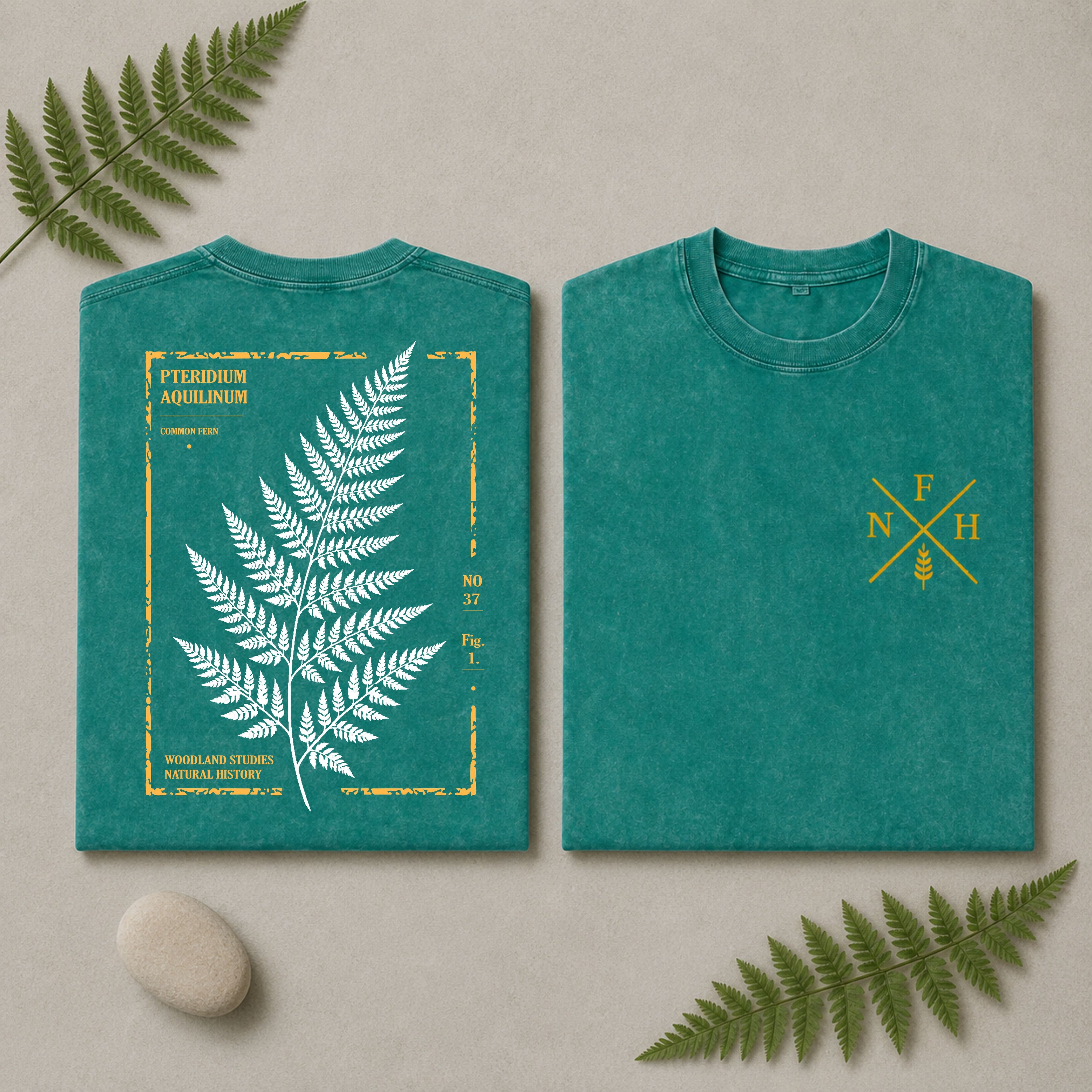 Two folded teal T-shirts displayed side by side on a light beige surface with decorative fern leaves and a small stone. The left shirt shows a large white fern illustration framed by distressed yellow text and border details, and the right shirt shows a small yellow monogram-style fern graphic on the chest.