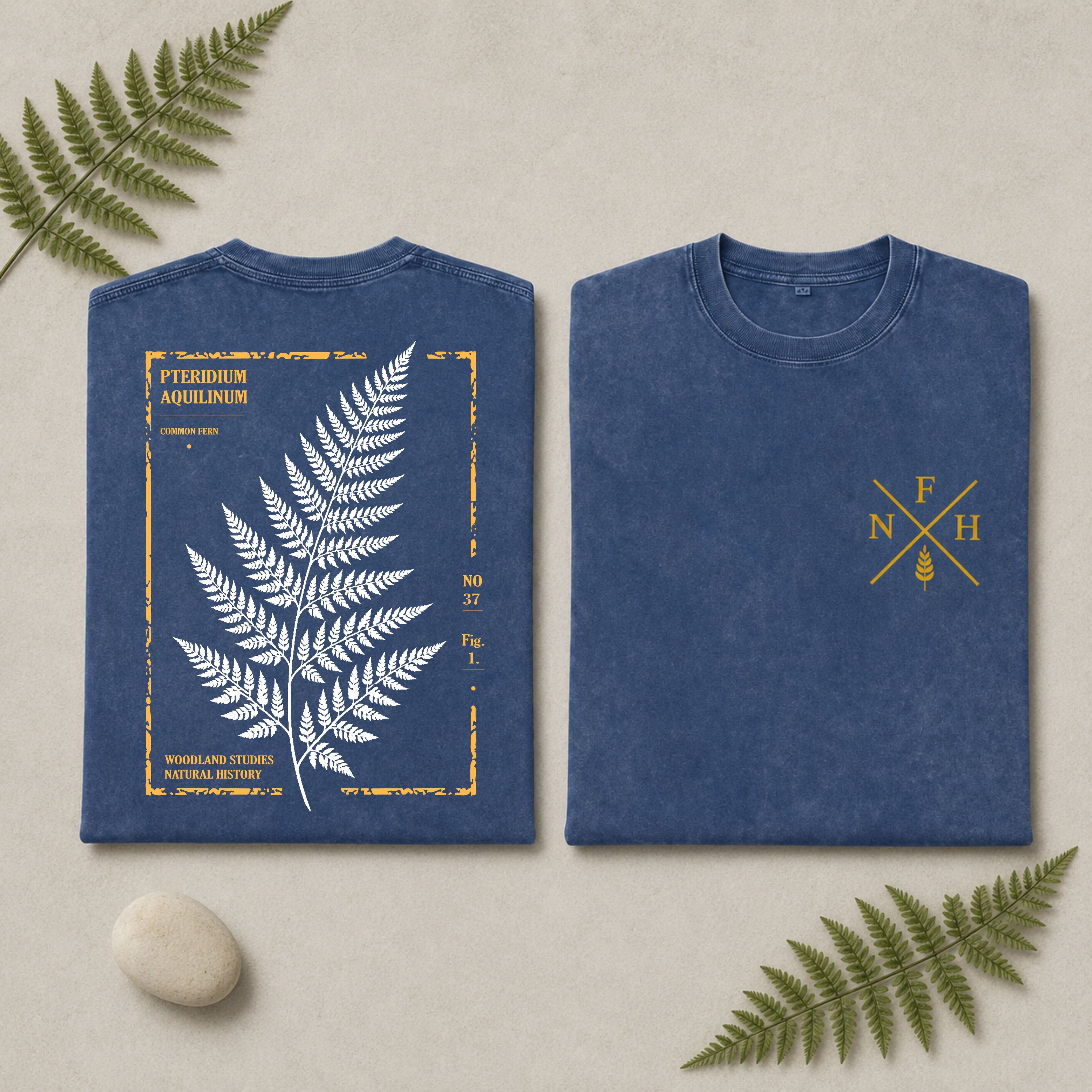 Two views of a blue crewneck t-shirt laid flat on a light beige background with decorative fern leaves and a small stone. The left view shows a large white fern illustration framed by a distressed gold border with text.