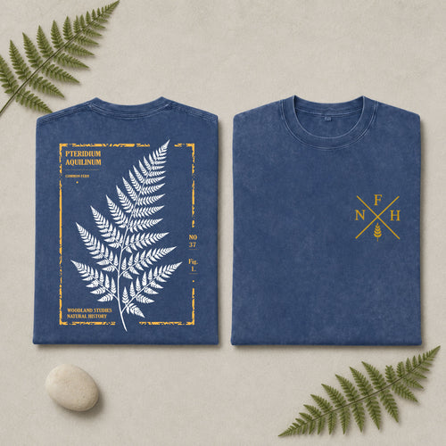 Two views of a blue crewneck t-shirt laid flat on a light beige background with decorative fern leaves and a small stone. The left view shows a large white fern illustration framed by a distressed gold border with text.