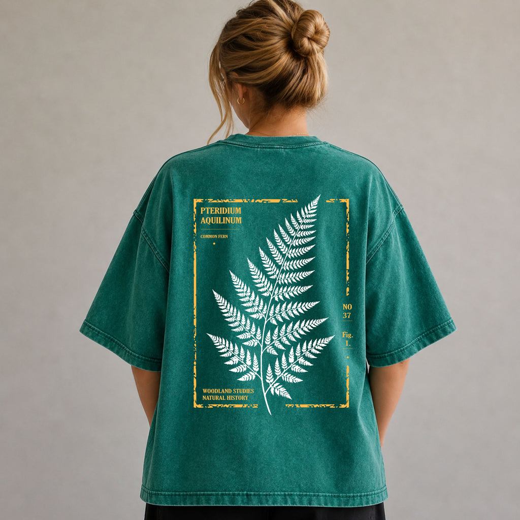 A woman viewed from behind wearing an oversized teal-green T-shirt with a large white fern illustration and yellow vintage-style text printed on the back, set against a plain light gray wall.