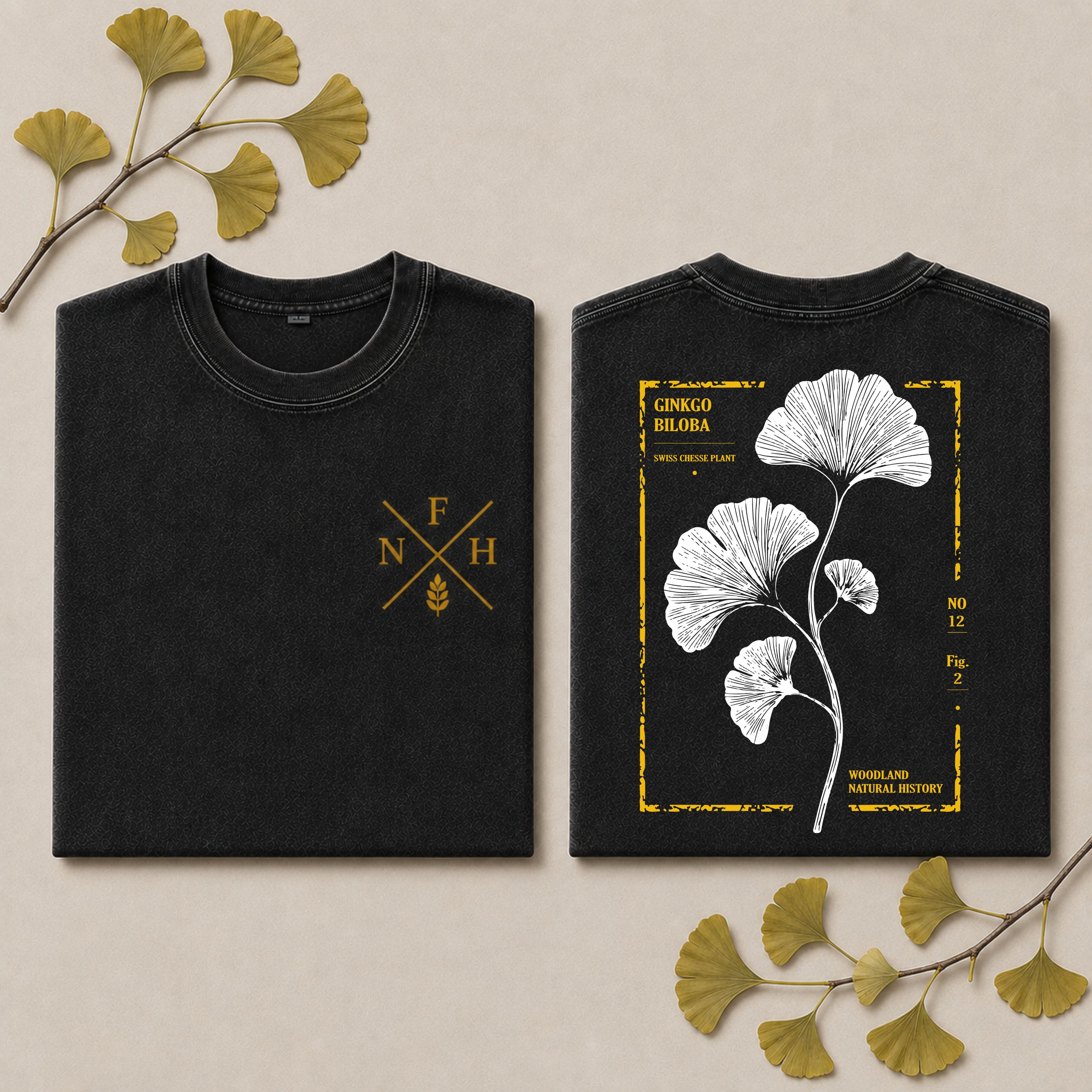 Two folded black T-shirts shown side by side on a light beige background with decorative ginkgo leaves. The left shirt has a small gold monogram-style chest print reading F N H, and the right shirt has a large white botanical illustration of ginkgo leaves framed by a distressed gold border with text including 'GINKGO BLADIA' and 'WILDERNESS NATURE HISTORY'.