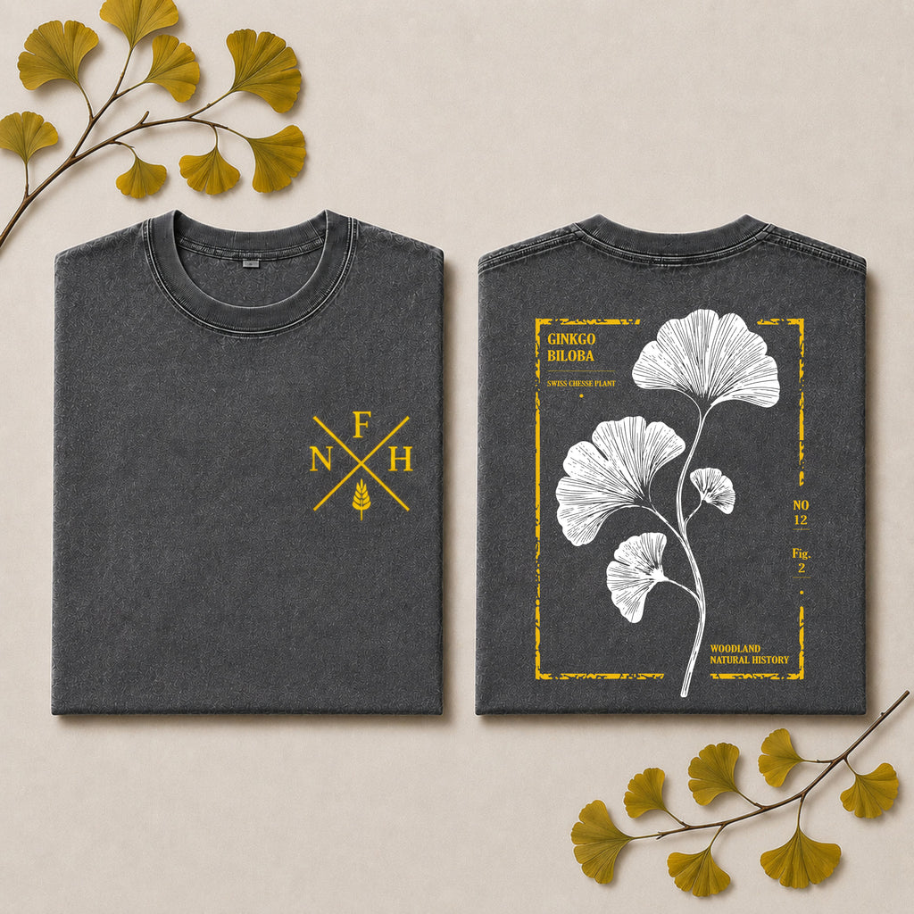 Two folded dark gray T-shirts shown side by side on a light beige background with decorative ginkgo leaves. The left shirt has a small yellow monogram-style chest print reading F N H with a ginkgo leaf motif.