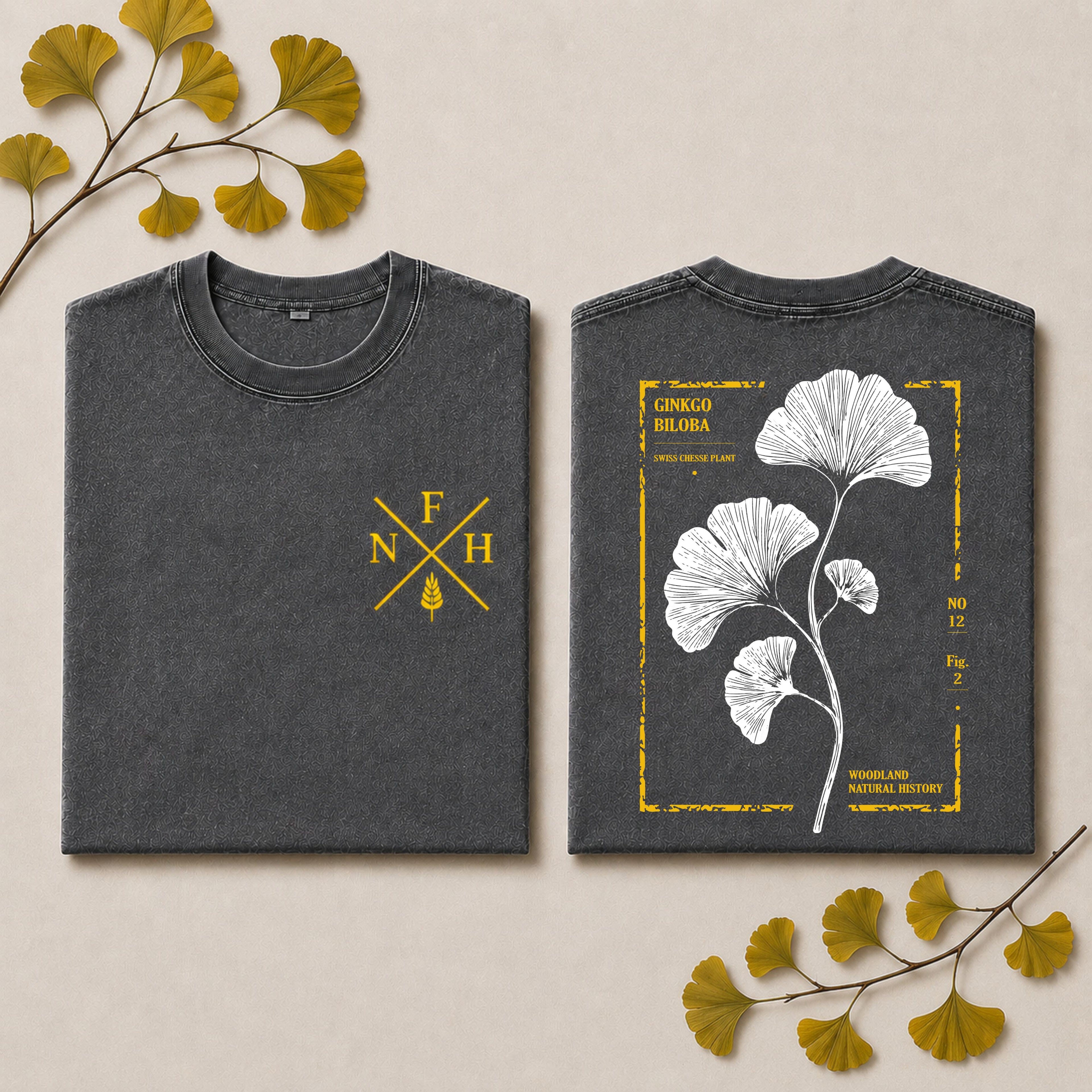 Two folded dark gray T-shirts shown side by side on a light beige background with decorative ginkgo leaves. The left shirt has a small yellow monogram-style chest print reading F N H with a ginkgo leaf motif.