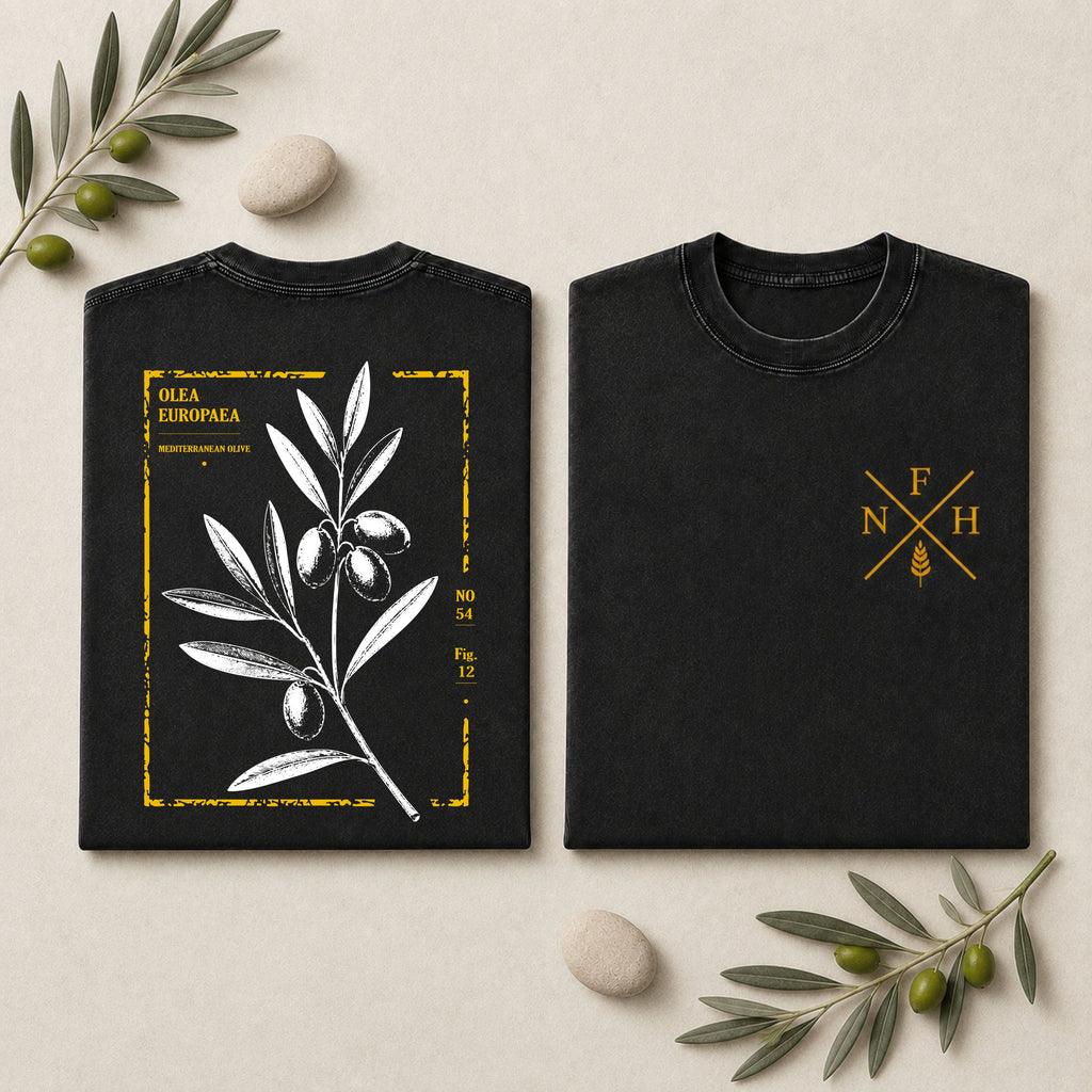 Two black short-sleeve T-shirts laid flat on a light beige surface with decorative olive branches and stones. The left shirt shows a large white olive branch illustration with a distressed gold border and text.