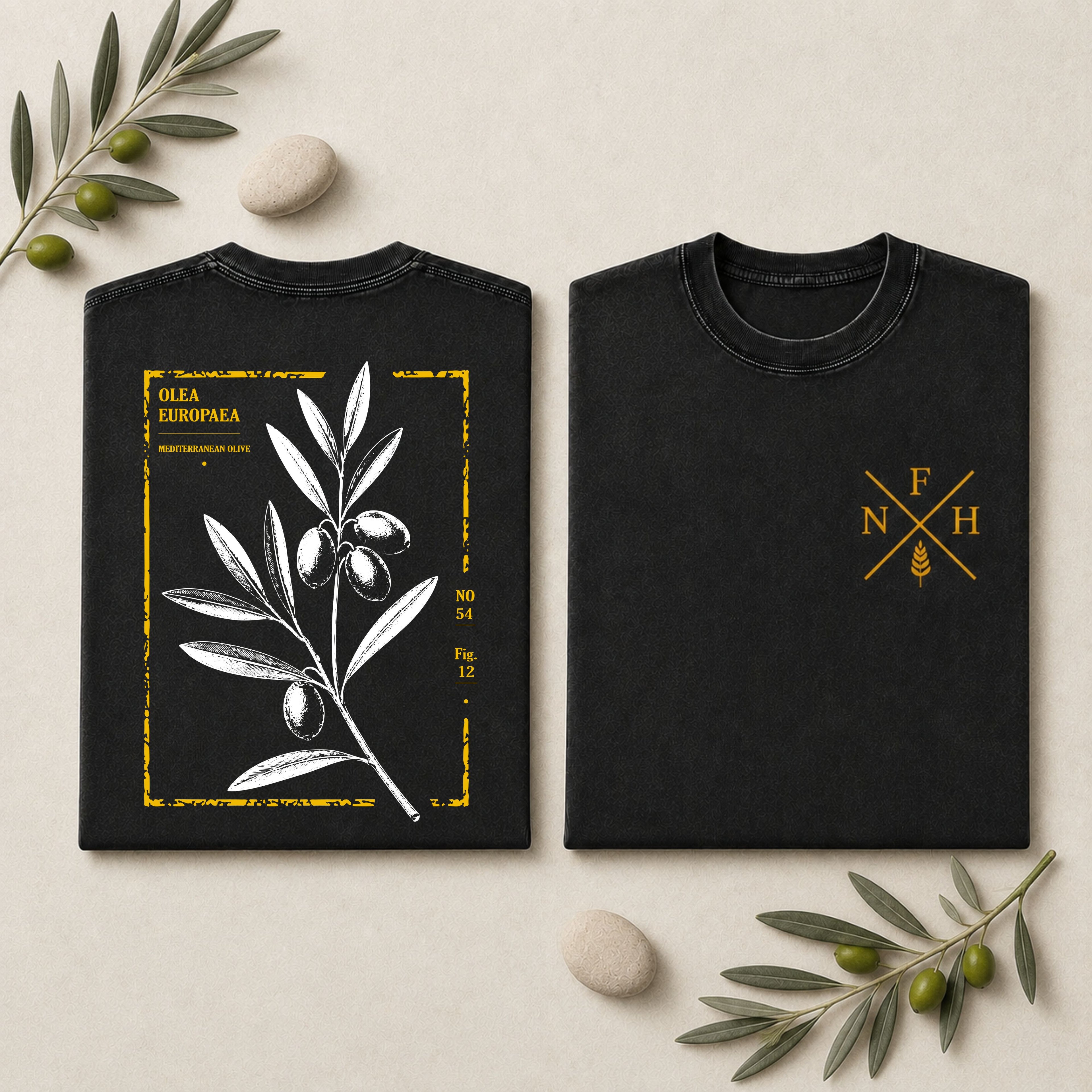 Two black short-sleeve T-shirts laid flat on a light beige surface with decorative olive branches and stones. The left shirt shows a large white olive branch illustration with a distressed gold border and text.