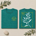 Two folded teal T-shirts shown side by side on a light beige background with olive branches and a stone. The left shirt has a small yellow chest logo with letters and wheat, and the right shirt has a large white olive branch illustration with yellow text and border.