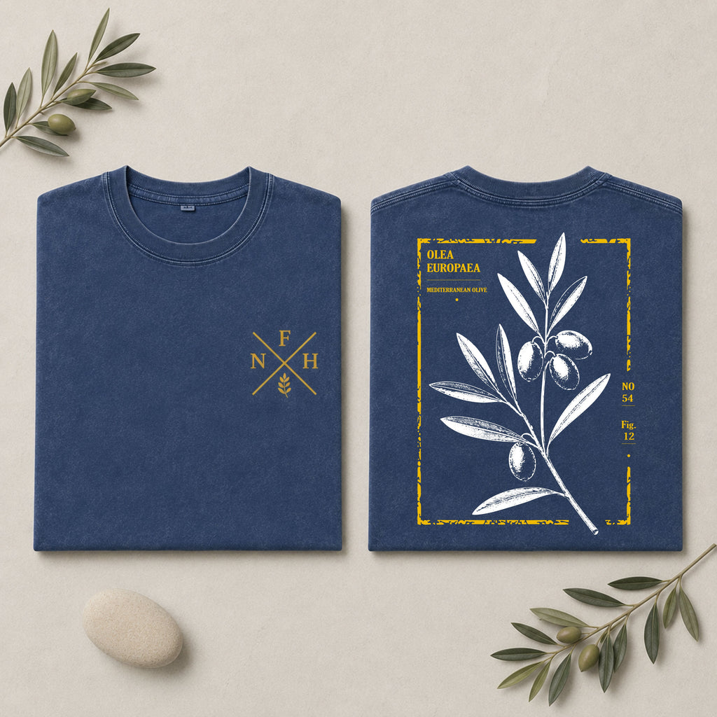 Two navy blue folded T-shirts shown on a light beige background with decorative olive branches and a stone. The left shirt has a small gold chest emblem, and the right shirt shows a larger back print with an olive branch illustration and yellow text reading 'OLIVA EUROPEA'.