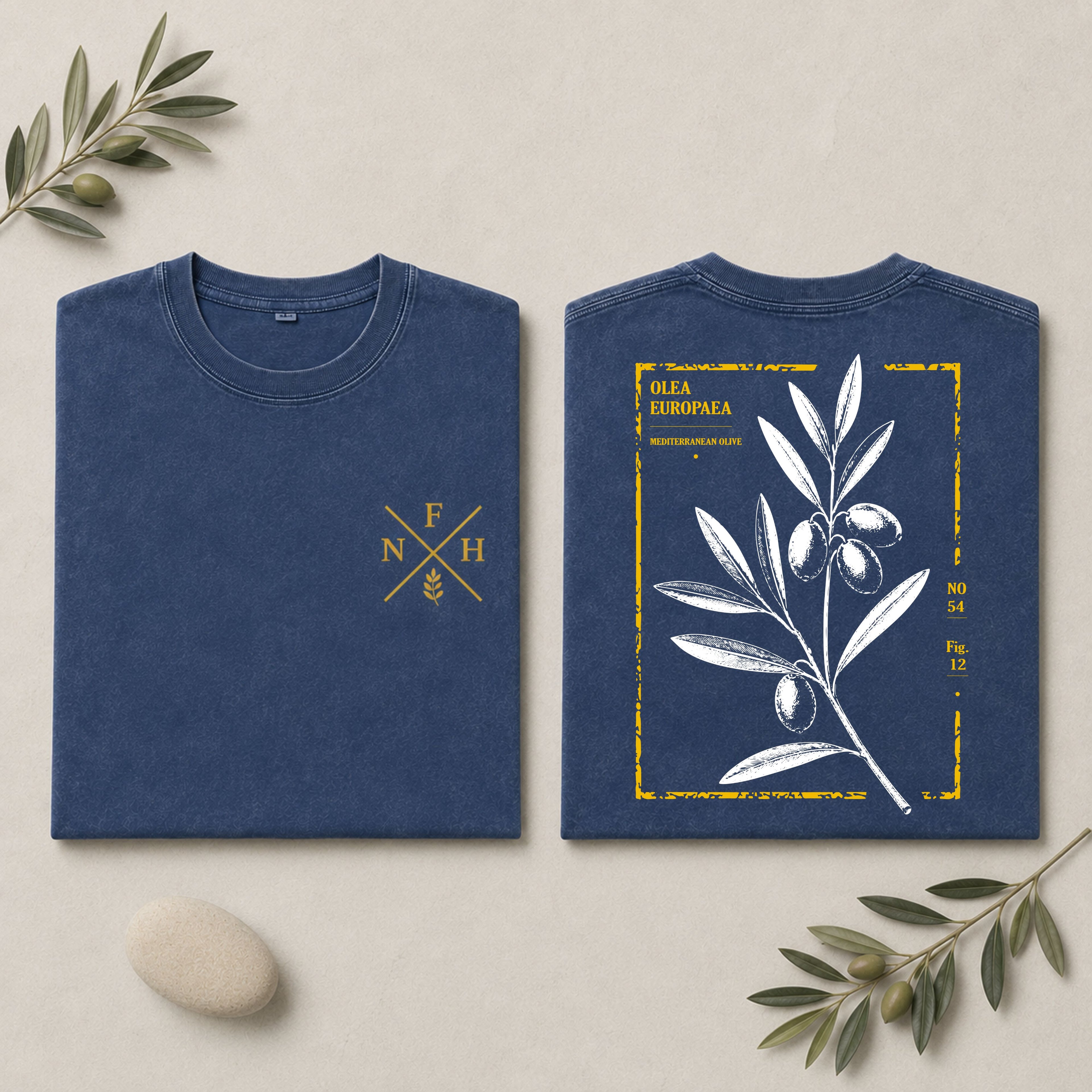 Two navy blue folded T-shirts shown on a light beige background with decorative olive branches and a stone. The left shirt has a small gold chest emblem, and the right shirt shows a larger back print with an olive branch illustration and yellow text reading 'OLIVA EUROPEA'.