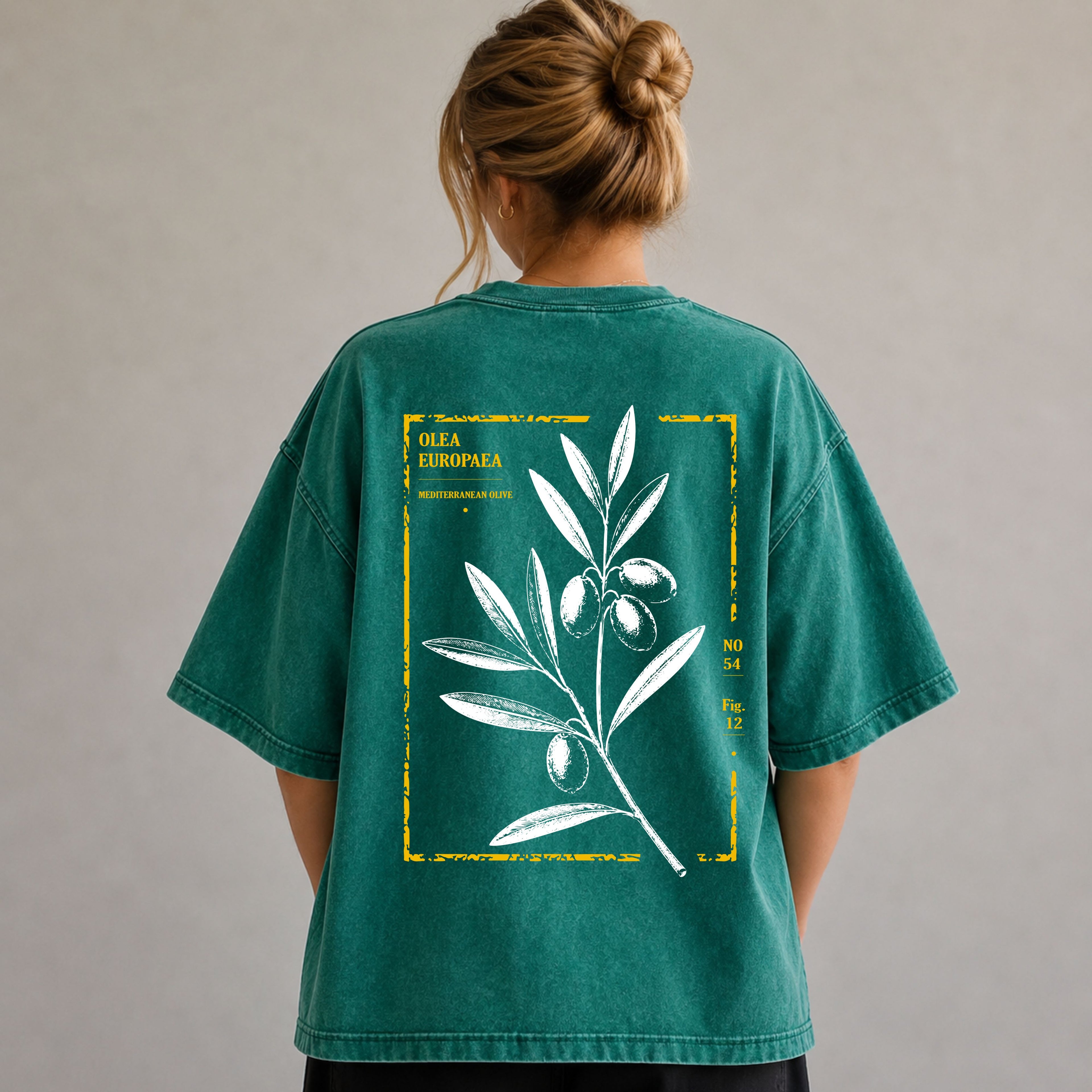 A woman viewed from behind wearing an oversized teal-green graphic T-shirt. The back of the shirt features a large white botanical illustration of an olive branch with green olives inside a distressed yellow rectangular border.